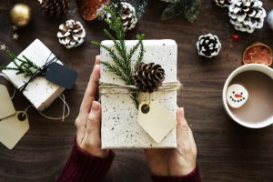 Two Hands holding a nicely wrapped gift tied with jute twine with a sprig of greens and a pine-cone on top. A mug full of cocoa decorated with a large marshmellow with a smiling face drawn on it.