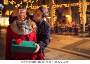 Man covering woman's eyes with one hand and holding a package wrapped in green giftwrap topped off with a red bow..