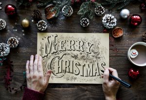 A Calligrapher holding a pen. With a beautiful hand-lettered plaque saying, "Merry Christmas".  It is placed on top of a background with snow-dusted pine cones and a mug of hot cocoa with a smiling faced marshmallow.